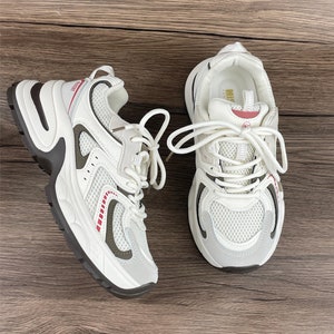 May include: A pair of white trainers with a mesh upper and a chunky sole. The trainers have a red and white stripe detail on the side and a white lace.
