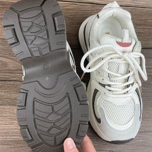 May include: White trainers with a brown rubber sole. The trainers have a mesh upper and a white lace-up closure.