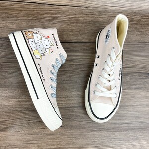 May include: A pair of light pink high-top canvas shoes with a black trim. The shoes have a cartoon design of cats stacked on top of each other with the text "meow" on the side.