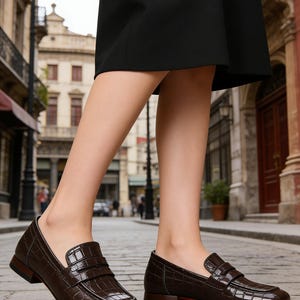 May include: Brown leather loafers with a crocodile texture. The shoes have a low heel and a strap across the top. The soles are a contrasting color. The image shows the shoes being worn on a cobblestone street.
