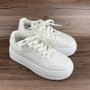 May include: A pair of white platform sneakers with white laces. The sneakers have a textured leather upper and a thick, multi-layered platform sole.