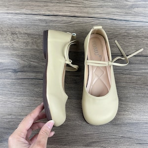 May include: A pair of light beige ballet flats with a brown sole and a tie closure. The flats have the word "TOMU" printed on the inside of the right shoe.