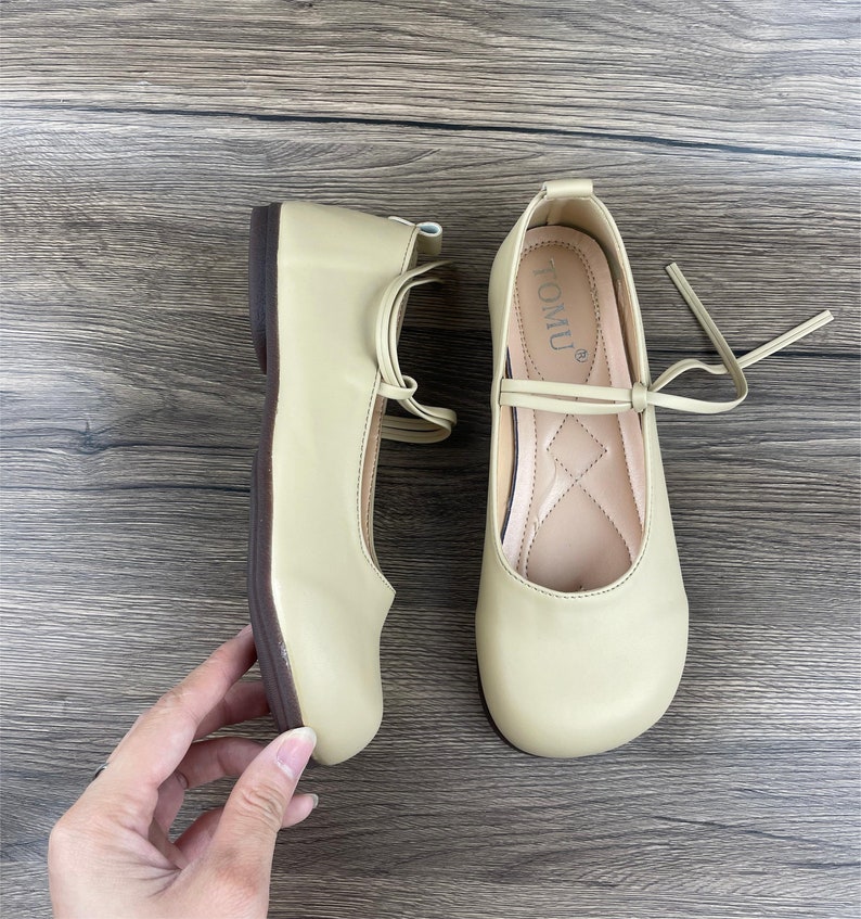 May include: A pair of light beige ballet flats with a brown sole and a tie closure. The flats have the word "TOMU" printed on the inside of the right shoe.