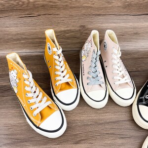 May include: Three pairs of high-top canvas shoes with cartoon cat designs and the text "Meow" on the side. One pair is yellow, one is beige, and one is black.