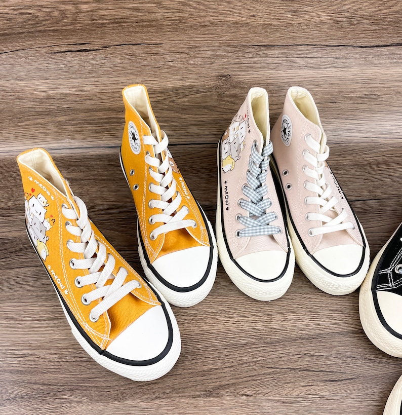 May include: Three pairs of high-top canvas shoes with cartoon cat designs and the text "Meow" on the side. One pair is yellow, one is beige, and one is black.