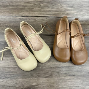 May include: Two pairs of beige and brown ballet flats with a ribbon tie. The shoes have a rounded toe and a low heel.