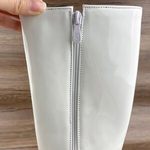 K&ouml;nnte beinhalten: Wei&szlig;e Lackleder-Stiefeletten, die bis zum Knie reichen, mit einem Rei&szlig;verschluss auf der R&uuml;ckseite. Der Rei&szlig;verschluss hat ein kleines wei&szlig;es Etikett mit dem Buchstaben "G" darauf.