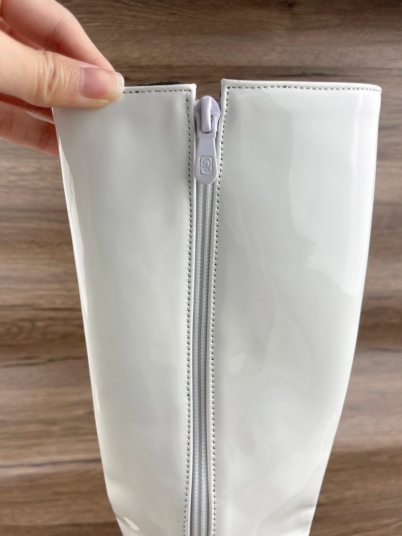 K&ouml;nnte beinhalten: Wei&szlig;e Lackleder-Stiefeletten, die bis zum Knie reichen, mit einem Rei&szlig;verschluss auf der R&uuml;ckseite. Der Rei&szlig;verschluss hat ein kleines wei&szlig;es Etikett mit dem Buchstaben "G" darauf.