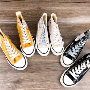 May include: Four pairs of high-top canvas sneakers with cartoon character designs. The sneakers are in yellow, beige, black, and white.
