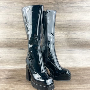 K&ouml;nnte beinhalten: Ein Paar schwarze Lackleder-Plateau-Stiefel mit einem dicken Absatz. Die Stiefel haben eine quadratische Spitze und einen Rei&szlig;verschluss an der Innenseite jedes Stiefels.