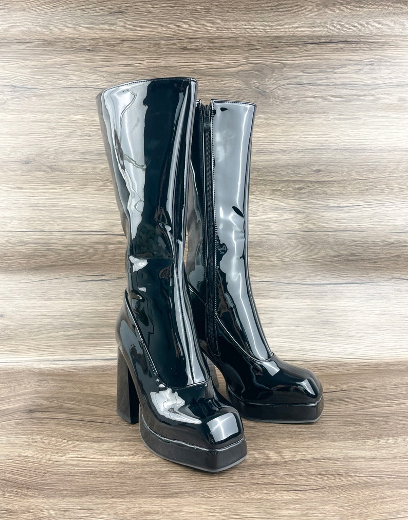 K&ouml;nnte beinhalten: Ein Paar schwarze Lackleder-Plateau-Stiefel mit einem dicken Absatz. Die Stiefel haben eine quadratische Spitze und einen Rei&szlig;verschluss an der Innenseite jedes Stiefels.