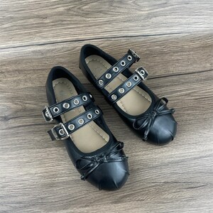 May include: A pair of black Mary Jane style flats with double straps, silver buckles, and decorative bows. The straps feature silver grommets. The shoes are displayed on a wooden surface.