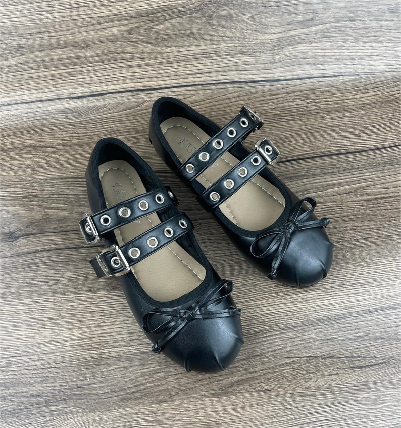 May include: A pair of black Mary Jane style flats with double straps, silver buckles, and decorative bows. The straps feature silver grommets. The shoes are displayed on a wooden surface.