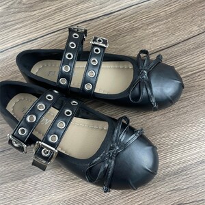May include: A pair of black ballet flats with double straps featuring silver grommets and buckles. Each shoe has a small bow on the toe. The shoes are displayed on a wooden surface.