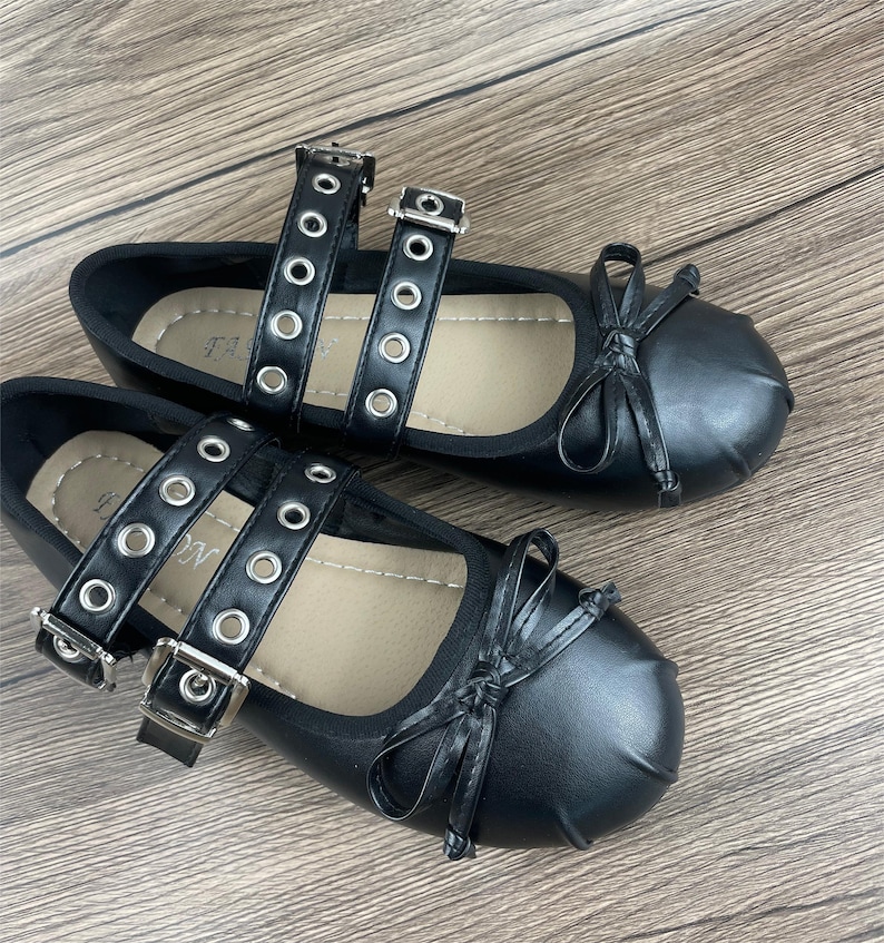 May include: A pair of black ballet flats with double straps featuring silver grommets and buckles. Each shoe has a small bow on the toe. The shoes are displayed on a wooden surface.
