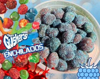 May include: A clear plastic container filled with blue and purple gummy candies, covered in a blue sugar coating. The candies are arranged in a single layer. The container is surrounded by red, green, and blue gummy candies. The text "Fruit Gushers Enchilados" is visible on the left side of the image.