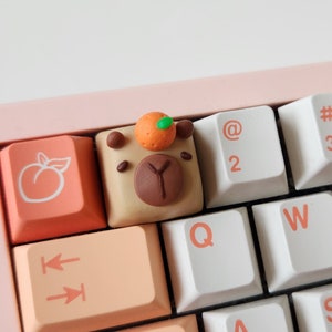 May include: A pink and white keyboard with custom keycaps. The keycaps are peach, brown, and white with peach and brown designs. The keyboard has the following keys: ←, @, #, S, 2, 3, 4, Q, W, E, R, A, S, D, Z, X, C.