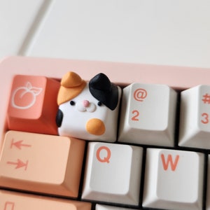 May include: A white keyboard with peach-colored keys and a cat-shaped keycap. The cat keycap is white with black ears, orange cheeks, and a black nose.