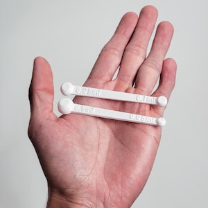 May include: Two white measuring spoons with rounded ends and printed measurements of 0.25ml, 0.5ml, 0.1ml, and 0.05ml. The spoons are held in an open hand against a plain background. The spoons are designed for precise measurements.