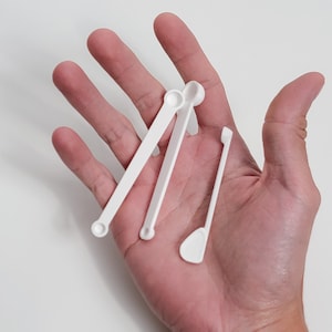 May include: A collection of white plastic measuring spoons and a small spatula are displayed. The spoons have different sizes and shapes, with a rounded scoop at the end of each. The spatula has a flat, rectangular head. The items are held in a hand.
