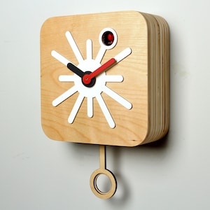 May include: A contemporary wooden wall clock in light brown. It has a white starburst design, black and red hands, and a circular pendulum. The clock measures approximately 25 cm wide and 30 cm tall.