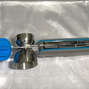 May include: A stainless steel cocktail jigger with a blue silicone end and a multi-tool. The jigger has two measuring cups. The multi-tool has a gray and blue handle. The items are on a white surface.