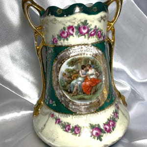 Könnte beinhalten: Eine antike Porzellanvase mit zwei verzierten goldenen Griffen. Die Vase ist mit einem grünen Hintergrund, goldenen Akzenten und einer zentralen Szene von zwei Frauen in einem Garten verziert. Die Szene ist von einem goldenen Kreis und einem Rand aus rosa Rosen umgeben.