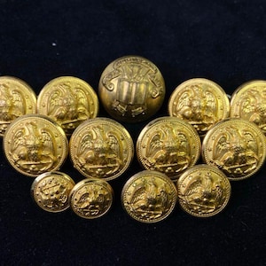 May include: A collection of antique gold-coloured buttons. The buttons feature an eagle design, with some having a shield and banner motif. The buttons vary in size and are arranged on a dark background.