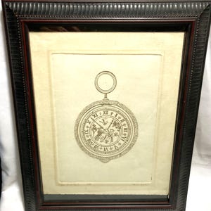 Pode incluir: Impressão emoldurada de um relógio de bolso antigo. O mostrador do relógio apresenta algarismos romanos e uma ilustração detalhada. A moldura é marrom escuro com uma borda decorativa. A impressão está em um fundo creme.