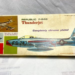 Puede incluir: Caja de modelo de avión Republic F-84G Thunderjet vintage. La caja presenta una ilustración detallada del avión cromado, con el texto "Thunderjet" y "FS-747". La caja también incluye una imagen de un modelo terminado.