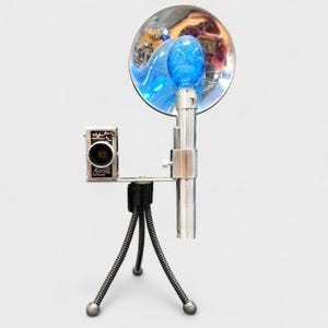 May include: A vintage camera with a silver body is mounted on a small tripod. A large, chrome reflector with a blue light bulb is attached to the camera. The camera and flash are set against a light gray background.