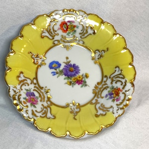 May include: A small, yellow porcelain plate with a white center and gold trim. The plate features a floral design with purple, blue, and yellow flowers.