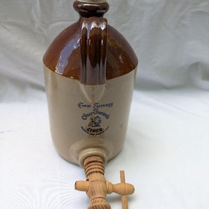 Könnte beinhalten: Ein Vintage-Steinzeug-Apfelwein-Krug mit braun glasierter Oberseite und Griff sowie einem beigefarbenen Sockel. Der Krug verfügt über einen Holzspund und einen braunen Verschluss. Der Krug trägt den blauen Text "Farm Scrumpy Churton's Cider".