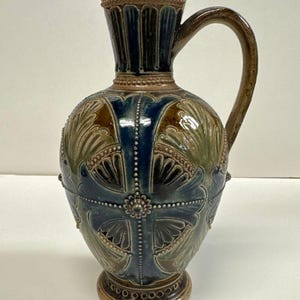 Könnte beinhalten: Dekorative Keramikkrug mit Henkel. Der Krug hat einen braunen Rand und Henkel, einen blauen Hals mit vertikalen Rillen und einen Körper, der mit blauen, braunen und grünen blattartigen Mustern verziert ist. Der Sockel ist braun.