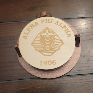Könnte beinhalten: Ein Satz von vier Untersetzern aus Holz mit braunem Sockel. Der oberste Untersetzer zeigt ein goldenes, graviertes Bild eines ägyptischen Pharaonenkopfes mit dem Text "ALPHA PHI ALPHA" und "1906" um das Bild herum.