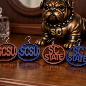 May include: Four circular drop earrings with silver-toned hooks. Two earrings are brown and two are blue. The brown and blue earrings feature the text SCSU in a cutout style, while the other brown and blue earrings feature the text SC STATE.