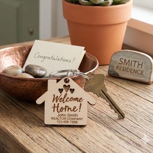 May include: A wooden house-shaped keychain with the words "Welcome Home!" and a realtor's contact information. The keychain is next to a key, a copper bowl with stones, a potted succulent, and a "Smith Residence" sign on a wooden table.
