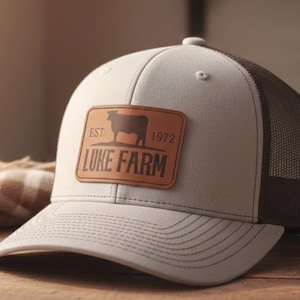 May include: A white and brown trucker hat with a leather patch. The patch features a cow silhouette and the text "LUKE FARM EST. 1972". The hat has a mesh back and a curved brim.