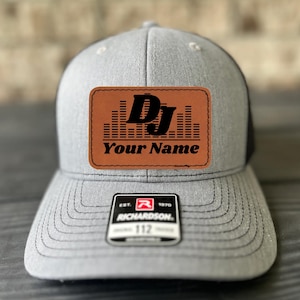 May include: A gray and black trucker hat with a brown leather patch that says "DJ" with sound waves and "Your Name". The hat has a black mesh back and a Richardson logo on the front.