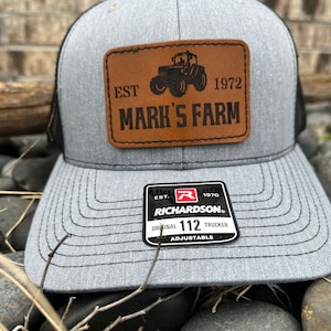 May include: Gray and black trucker hat with a brown leather patch that says "EST. 1972 MARK'S FARM" and a black tractor image. The patch is on the front of the hat. The hat has a black mesh back and a black adjustable strap with a black label that says "RICHARDSON® ORIGINAL 112 TRUCKER ADJUSTABLE".