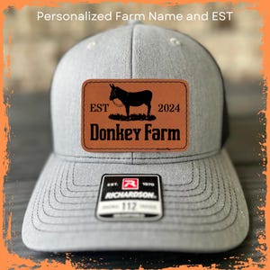 May include: A gray and black trucker hat with a brown leather patch that says "EST. 2024 Donkey Farm". The patch features a silhouette of a donkey. The hat also has a black and white Richardson logo.