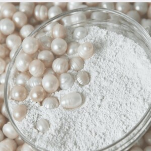 May include: A glass bowl filled with white powder and surrounded by white pearls. The pearls are arranged in a circle around the bowl.