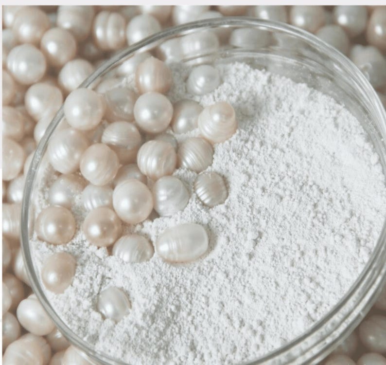 May include: A glass bowl filled with white powder and surrounded by white pearls. The pearls are arranged in a circle around the bowl.