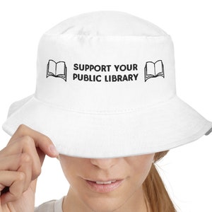 Peut inclure: Chapeau bob blanc avec un motif brodé noir qui dit "Support Your Public Library" avec une icône de livre ouvert de chaque côté du texte.