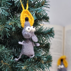 May include: A gray crocheted mouse ornament with large white eyes and a yellow crocheted star on top. The ornament is hanging on a green artificial Christmas tree.