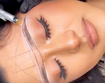 May include: A woman with her eyes closed is having her eyebrows shaped. White lines are drawn on her face to guide the eyebrow shaping process. A gloved hand holds a brush and applies a white substance to the eyebrow area.