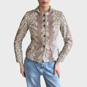 Op de afbeelding: Een blazer met slangenpatroon in beige, lichtbeige en roze tinten. De jas heeft een kraag, drie zwarte knopen en twee kleine zakken. De blazer met lange mouwen wordt gedragen met een blauwe spijkerbroek.