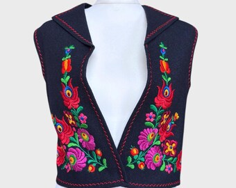 Pode incluir: Um colete preto com gola de xale, apresentando bordados florais vibrantes em vermelho, rosa, verde e amarelo. As bordas são adornadas com costuras vermelhas. O colete é exibido em um manequim branco.