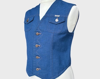 Op de afbeelding: Een blauw spijkergoed vest met twee borstzakken en een V-hals. Het vest heeft koperkleurige knopen en stiksels. Het vest wordt getoond op een witte mannequin.