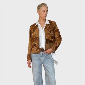 Peut inclure: Veste marron en patchwork avec un col. La veste présente une variété de tons et de textures marron. La personne porte une chemise blanche à col et un jean bleu clair.